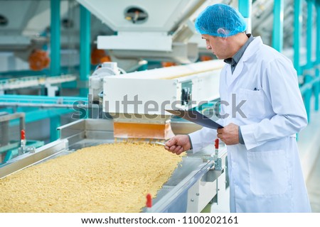 Side view portrait of senior factory worker doing  production quality inspection in food industry holding clipboard standing by conveyor belt, copy space