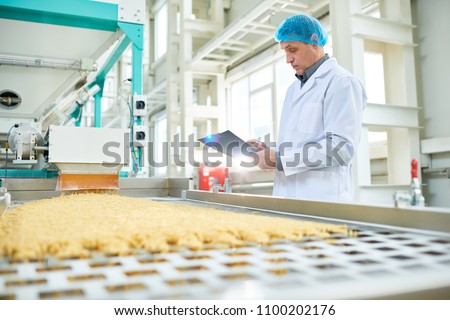 Side view portrait of senior factory worker  in food industry holding clipboard standing by conveyor belt, copy space