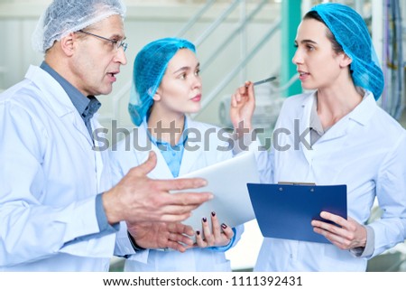 Waist up portrait of two young women asking questions to senior factory worker during training seminar for new specialists standing in clean workshop of modern plant.