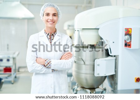 Beautiful confectionery factory worker standing in white coat with arms crossed smiling and looking at camera.