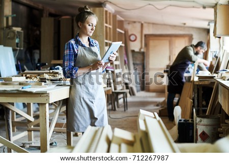 Full length portrait of concentrated young craftswoman in apron standing at spacious workshop and using digital tablet