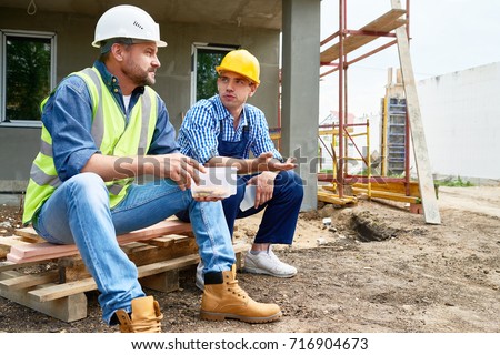 Two workers wearing protective helmets taking break from work and enjoying lunch while sitting outdoors, unfinished building on background