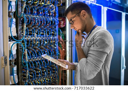 Side view portrait of young network engineer looking at digital tablet thoughtfully while working with servers in data center