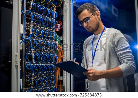 Portrait of young network engineer standing by server cabinet while working with supercomputer in data center and holding clipboard