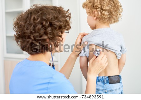 Portrait of female doctor listening to childs breathing from the back using stethoscope to check for symptoms of bronchitis or pneumonia