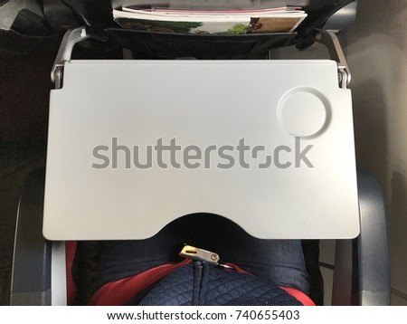 Passenger who wears blue jeans is fastening  red seat belt in airplane cabin. There are tray table and seat belt for passenger in each chair on the plane.