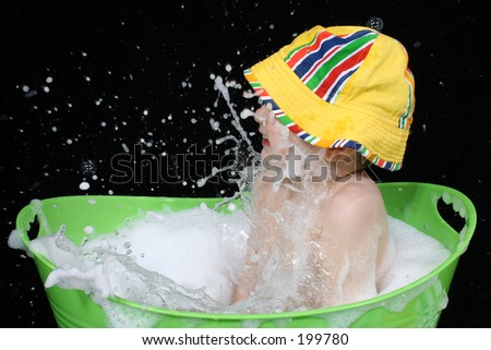 Two year old boy in a green tub of bubble water wearing a beach hat.