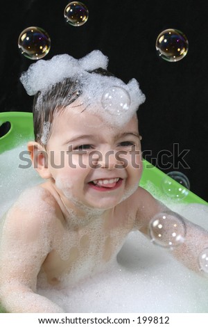 Two year old boy in a green tub of bubble water with bubbles on his head.