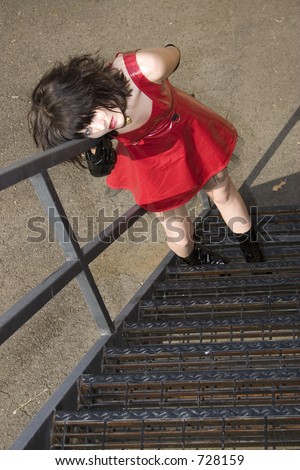 Beautiful Young Woman On Fire Escape. Wearing red vinyl dress, black boots and lock dog collar.