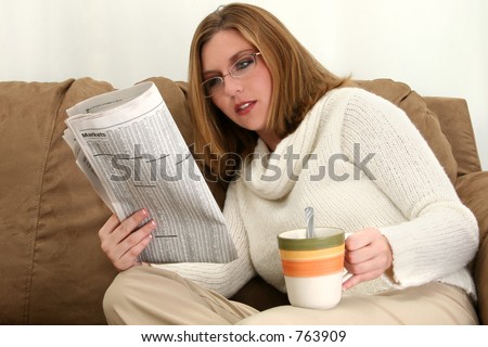 Beautiful young woman wearing sweater and glasses. Sitting on couch with cup of coffee reading newspaper.