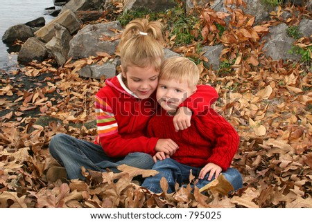 Brother and sister sitting in fall leaves hugging by the lake.