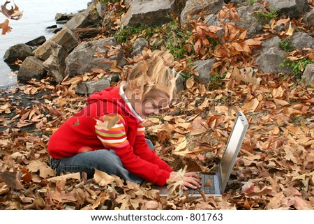 Girl on laptop in a pile of leaves.  Leaves falling on her.