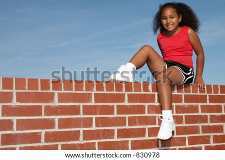 Beautiful seven year old African American girl sitting on brick wall against blue sky.