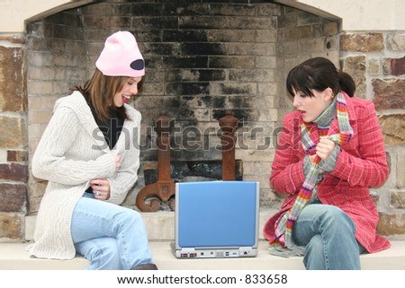 Two beautiful young woman (hispanic and caucasian) sitting outside by the fireplace with laptop.  Wearing winter clothes.