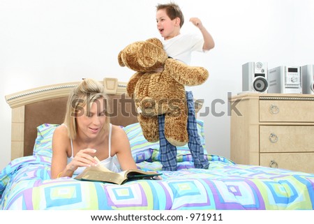 Mom reading book to son while he plays with giant stuffed bear in bed.