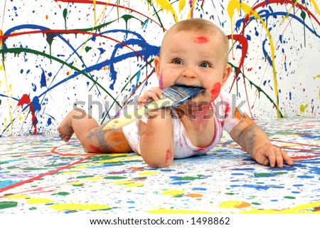Beautiful baby covered in bright paint with paint brush in mouth.