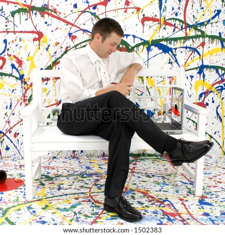 Attractive young business man sitting on white bench on paint splattered background.  Looking at laptop.  Full body.