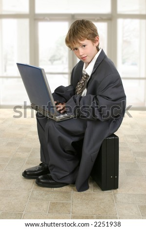 Adorable eight year old boy in father\'s business suit sitting on briefcase with laptop computer.