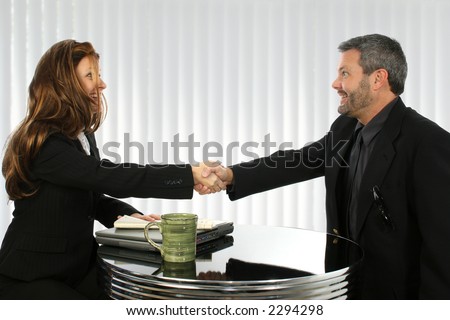 Business man and woman greeting at table with laptop and newspaper.