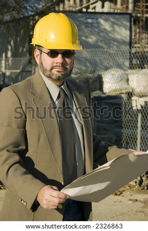 Man in suit at construction site holding bluerints and wearing yellow hardhat.
