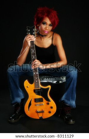 Beautiful young African American woman sitting on amp holding electric guitar over black.