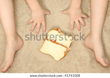 Young child's hands and feet in sand with peanut butter jelly sandwich.
