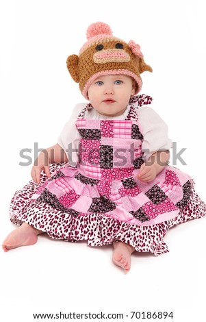 Attractive 6 month old baby girl in colorful dress, barefoot, and monkey cap over white.