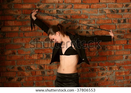 Girl in black suit tuck necktie above her head stands at brickwall.