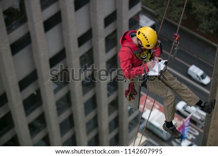 Wide angle pic of male rope access inspector worker working at height and abseiling down, hanging on rope in fall arrest position at high rise building written maintenance report,  Sydney, Australia