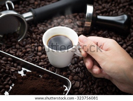 Fresh espresso. Hand grabbing an ear cup of of hot espresso coffee also see an espresso machine group head, coffee tamper and ground coffee lay on many roasted coffee beans.