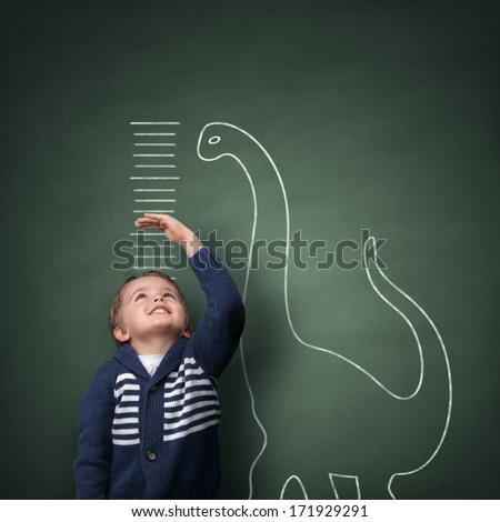 Young Boy Measuring His Growth In Height Against A Blackboard With Chalk Dinosaur Scale