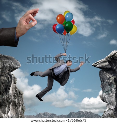 Businessman holding on to balloons above a cliff trying to escape hand bursting his balloon and bridge the gap concept for business adversity, risk, challenge or bullying