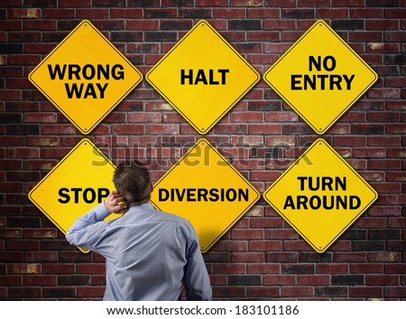 Businessman going the wrong way in front of a brick wall with stop, wrong way, halt, no entry, diversion and turn around road signs concept for blocked direction in business