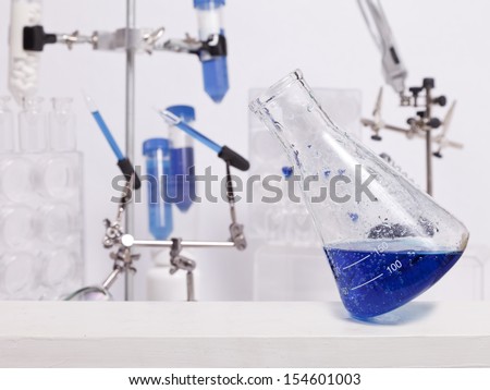 A science beaker with a blue liquid balances on its edge in a lab with other science equipment.