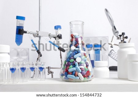 A science beaker full of different colored and shaped pills sits in a lab with other science equipment.