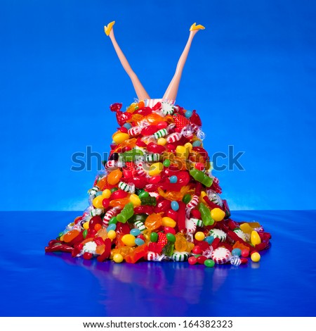 Legs and high heels from a generic children doll stick out of a pile of candy against a blue background.