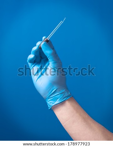 A male caucasian arm wearing a blue latex glove holding an eye dropper against a blue background.