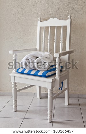Two rolled up white  hotel towels sit on a white chair on a white tiled floor.