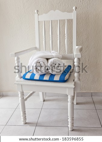Two rolled up white  hotel towels sit on a white chair on a white tiled floor.