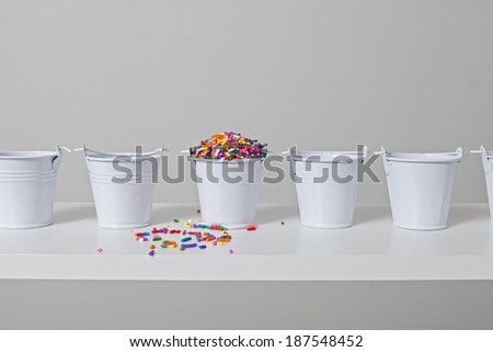 One small white bucket is filled to the brim with colorful cake decorating sprinkles and a few have spilled over.