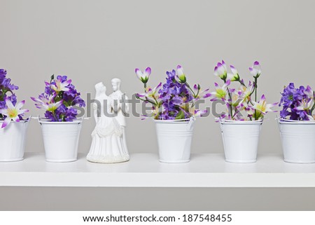 A bride and groom cake topper in a row with several small bouquets of purple flowers in tiny pails against a grey toned background.