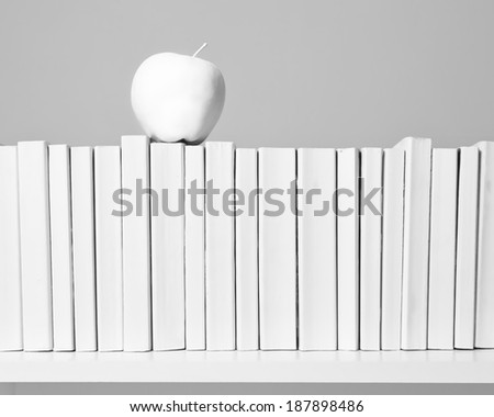 A row of all white books sit on a white shelf with a white apple in front of an off white background.