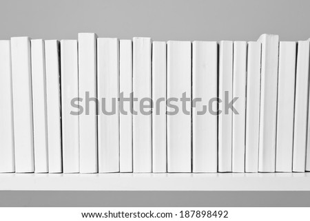A row of all white books sit on a white shelf in front of an off white background.