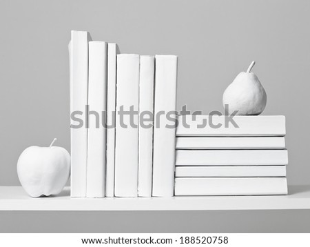 An arrangement of all white books sit on a white shelf with a white apple against a light grey background.