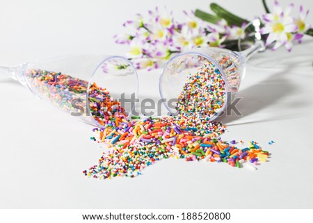 Two champagne glasses full of cake sprinkles lie on their sides and spill the multiple colored cake decorations over a white background.