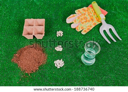 An arrangement of supplies needed to plant seeds including gardening gloves, a trowel, some dirt, water and seeds all against a green background.