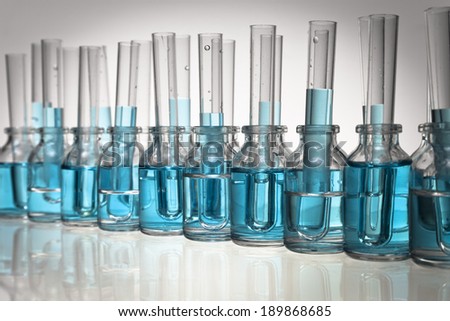 An arrangement of glass science vials and test tubes with some filled with a light blue liquid on a glass tabletop in a diagonal with vignette.