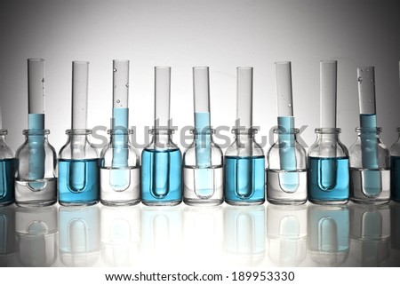 An arrangement of glass science vials and test tubes with some filled with a light blue liquid on a glass tabletop with a vignette.