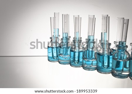 An arrangement of glass science vials and test tubes with some filled with a light blue liquid on a glass tabletop in a spotlight against a white background.