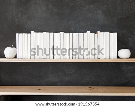 White Books Shelf on Blackboard. A row of all white books sit on the top shelf in front of a chalkboard with two white apples.